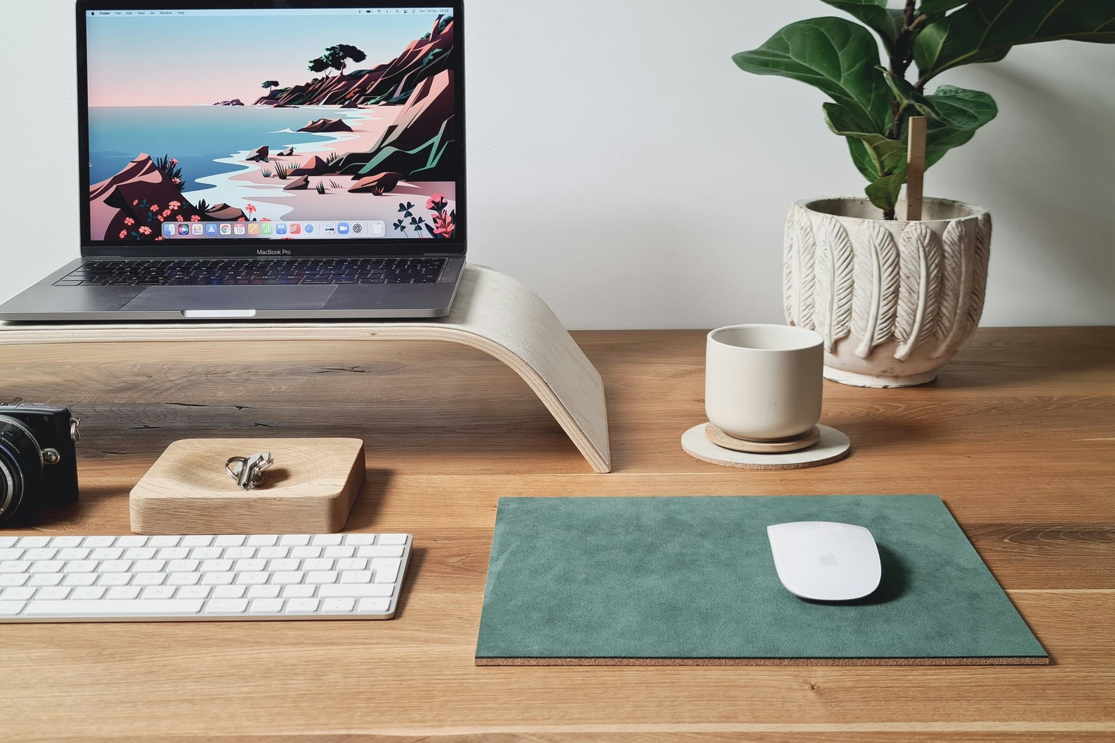 A MacBook Pro elevated on a white curved laptop stand on a wooden desk, with a plant, coffee cup, and keyboard.